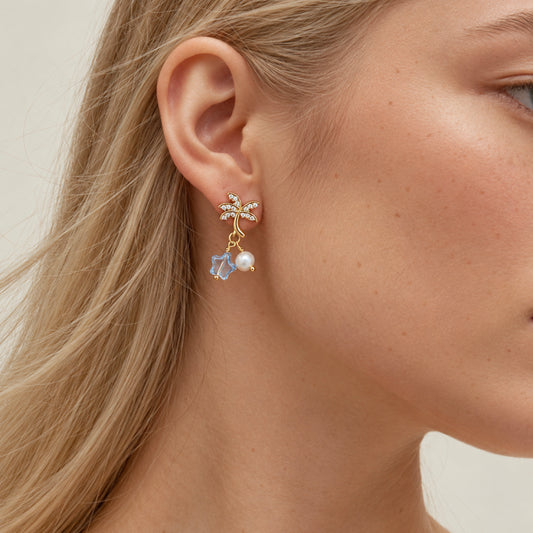 Starry Coconut Pearl Earrings by NINEMOO worn on the ear, with gold palm tree, blue star and white freshwater pearl.